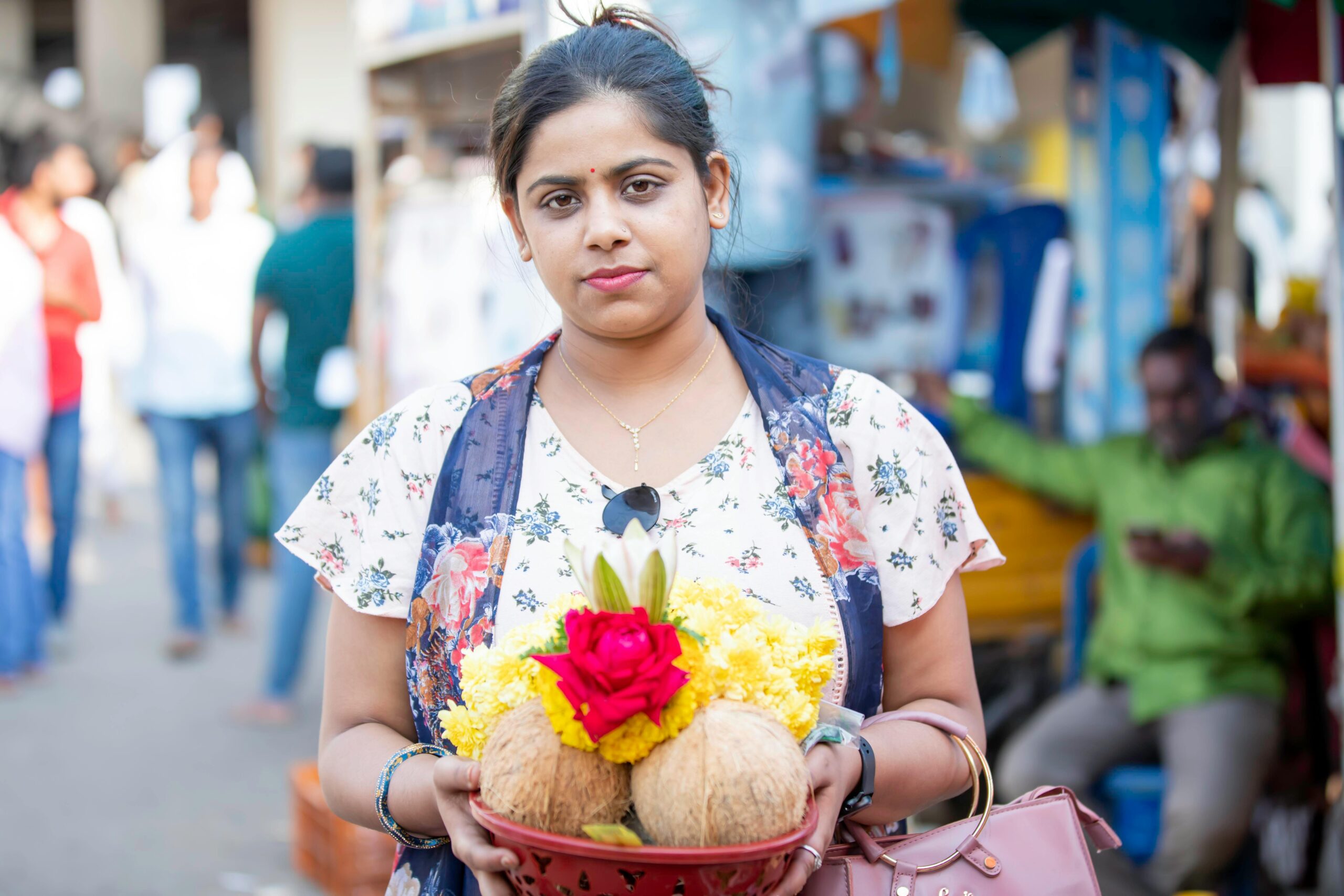 A woman in a floral dress holds colorful traditional offerings at a bustling Indian market.